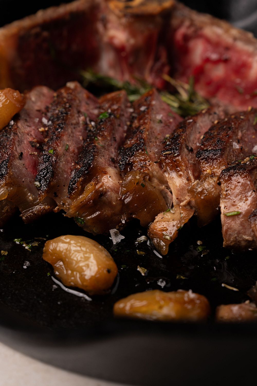 Sliced NY strip steak with seared crust, rosemary garnish, and roasted potatoes on dark plate.