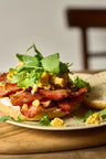 Sliced Ibérico coppa bacon layered on toasted bread with scrambled eggs, microgreens, and pea shoots on a white plate.