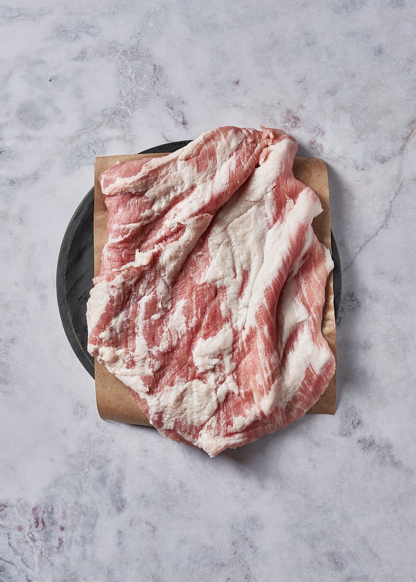 Raw Ibérico pork belly steak with marbled fat and meat on kraft paper and black plate.