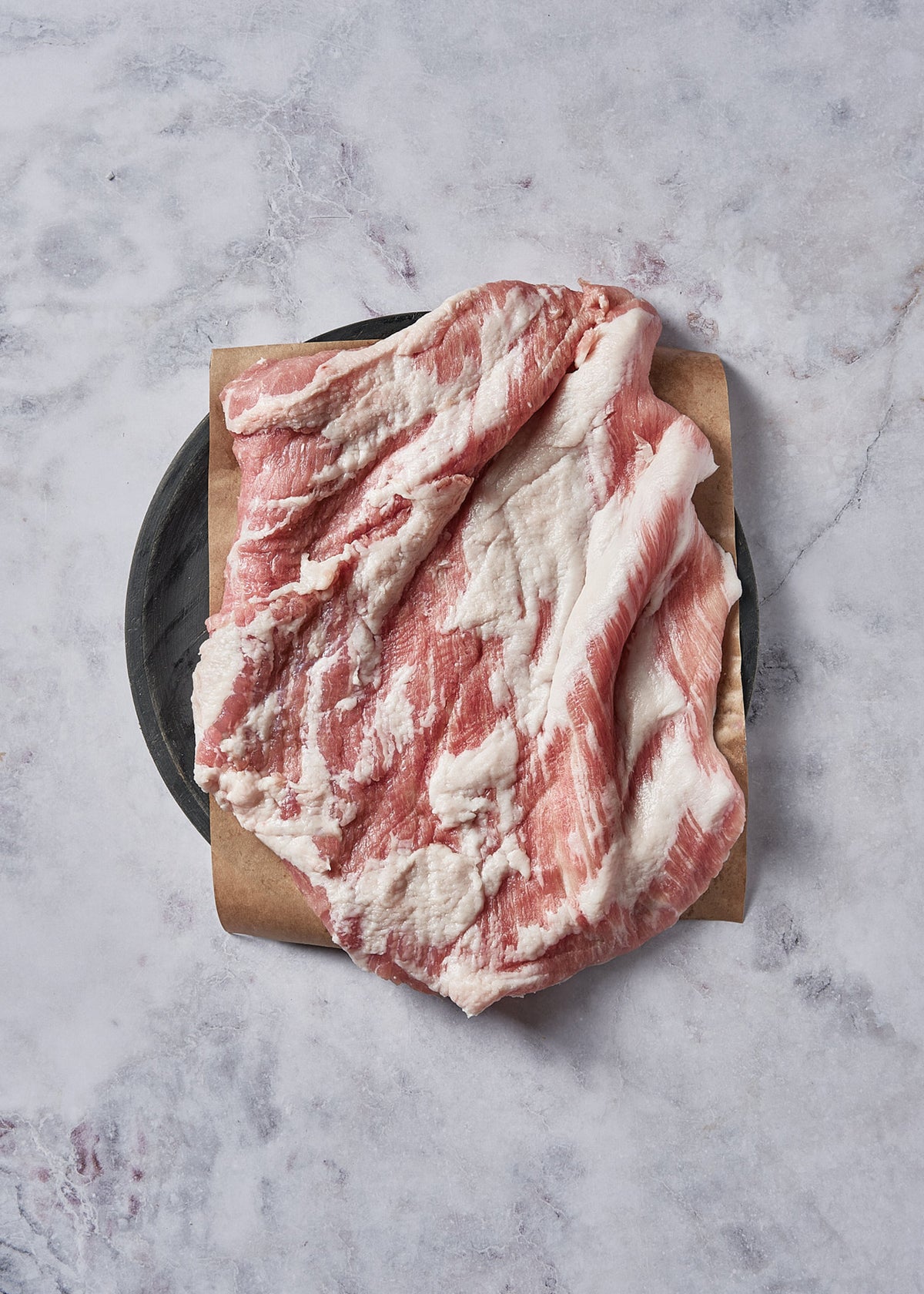 Raw Ibérico pork belly steak with marbled fat and meat on kraft paper and black plate.