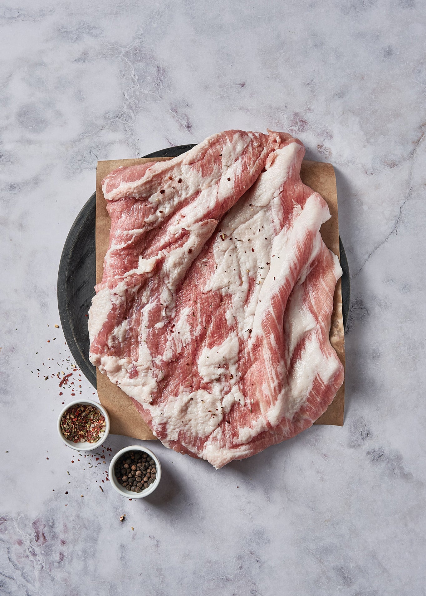 Sliced cured meat with marbled pink and white coloring on kraft paper with small bowls of peppercorns and seasoning blend.