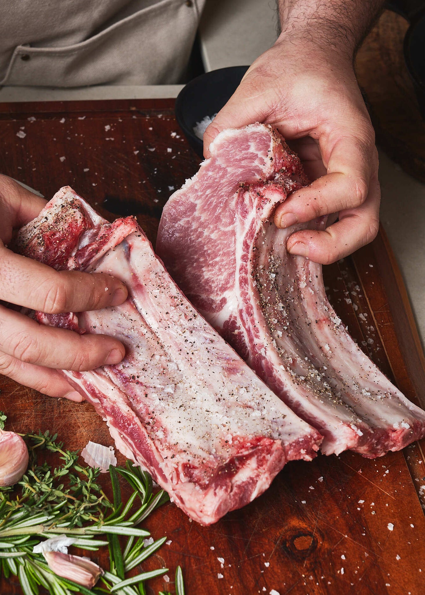 Four-rib Ibérico pork chops with seasoning held in hands over wooden board with fresh rosemary.