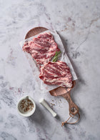 Two raw Ibérico short ribs with marbling on white paper and wooden board, garnished with rosemary.