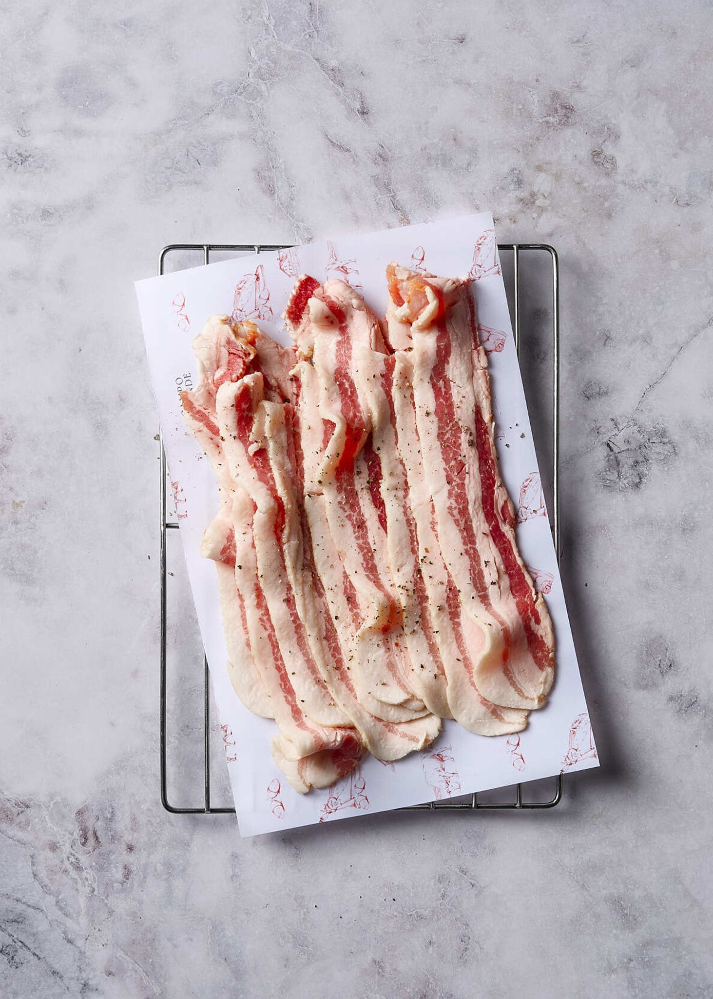 Sliced Ibérico bacon strips with red marbling on white paper atop a metal rack.