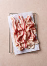 Sliced cured bacon with red and white marbling arranged on white paper with decorative pig illustrations.