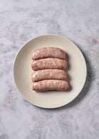 Four raw pork sausages arranged on a cream-colored plate against a marble surface.