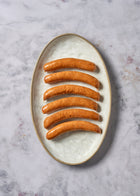 Six cooked sausages arranged on a cream-colored ceramic plate against a marble surface.