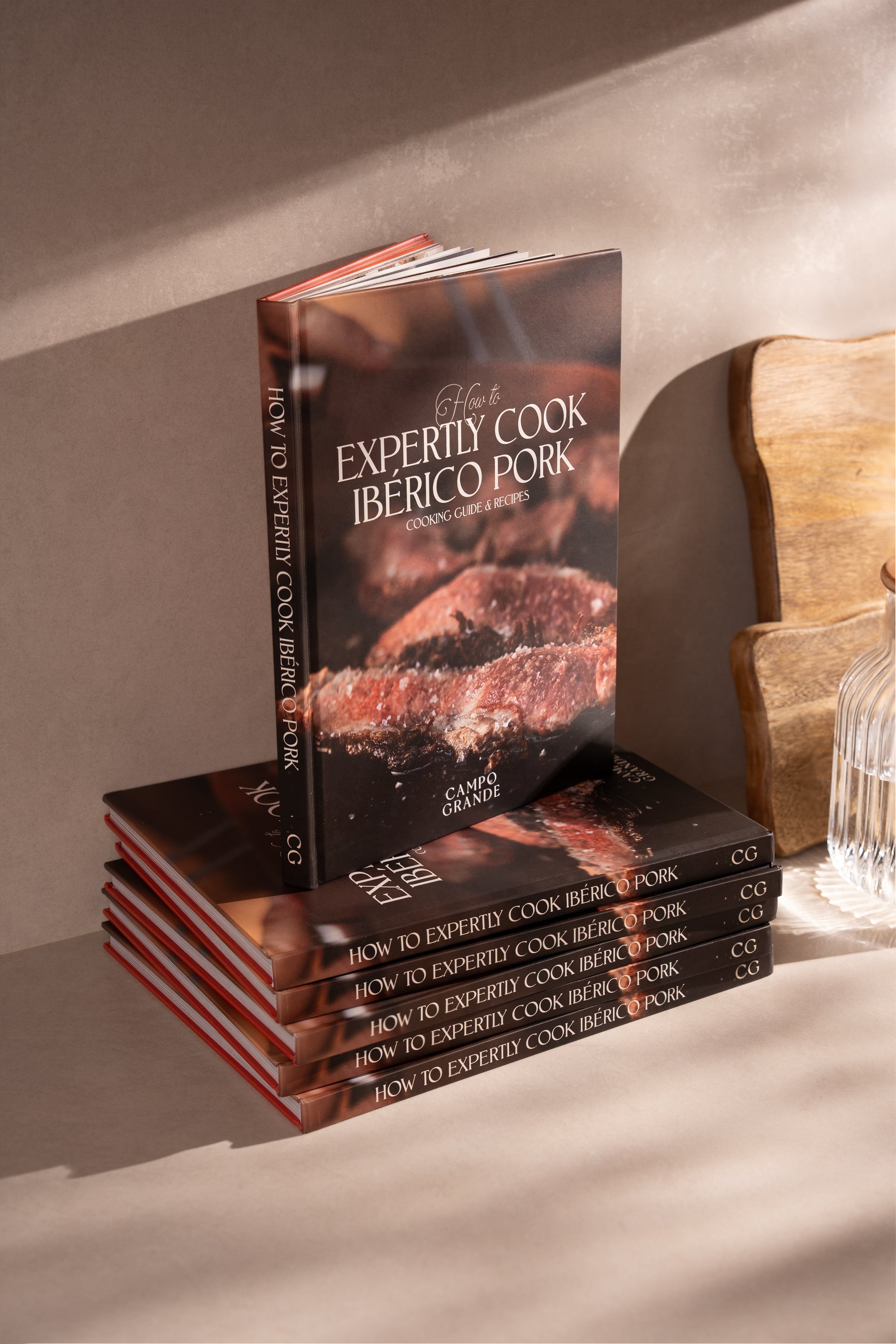 Stack of hardcover cookbooks titled "How to Expertly Cook Ibérico Pork" with dark cover featuring cooked meat, displayed next