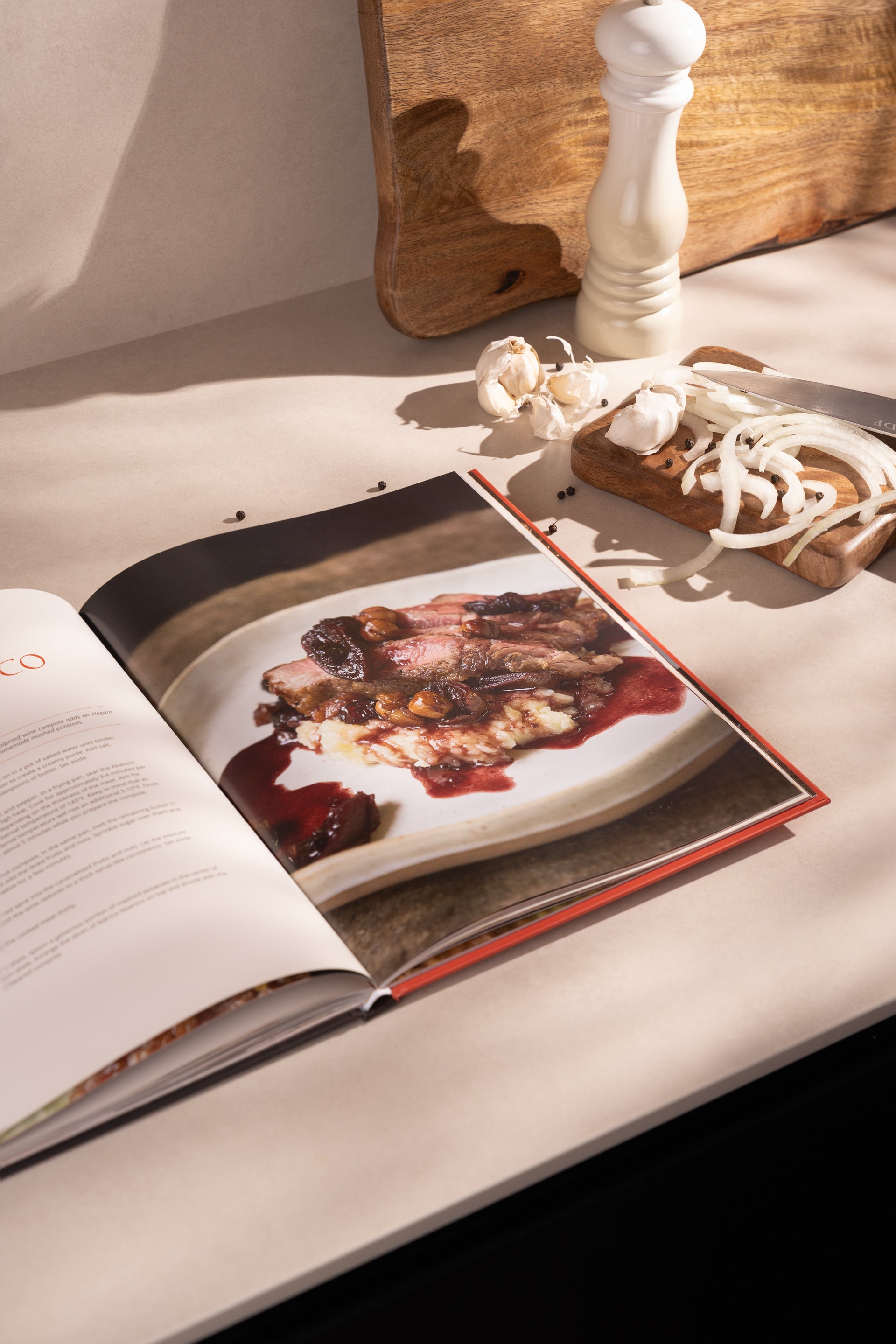 Open hardcover cookbook displaying cooked pork dish with red sauce, surrounded by wooden cutting board, white pepper mill, an