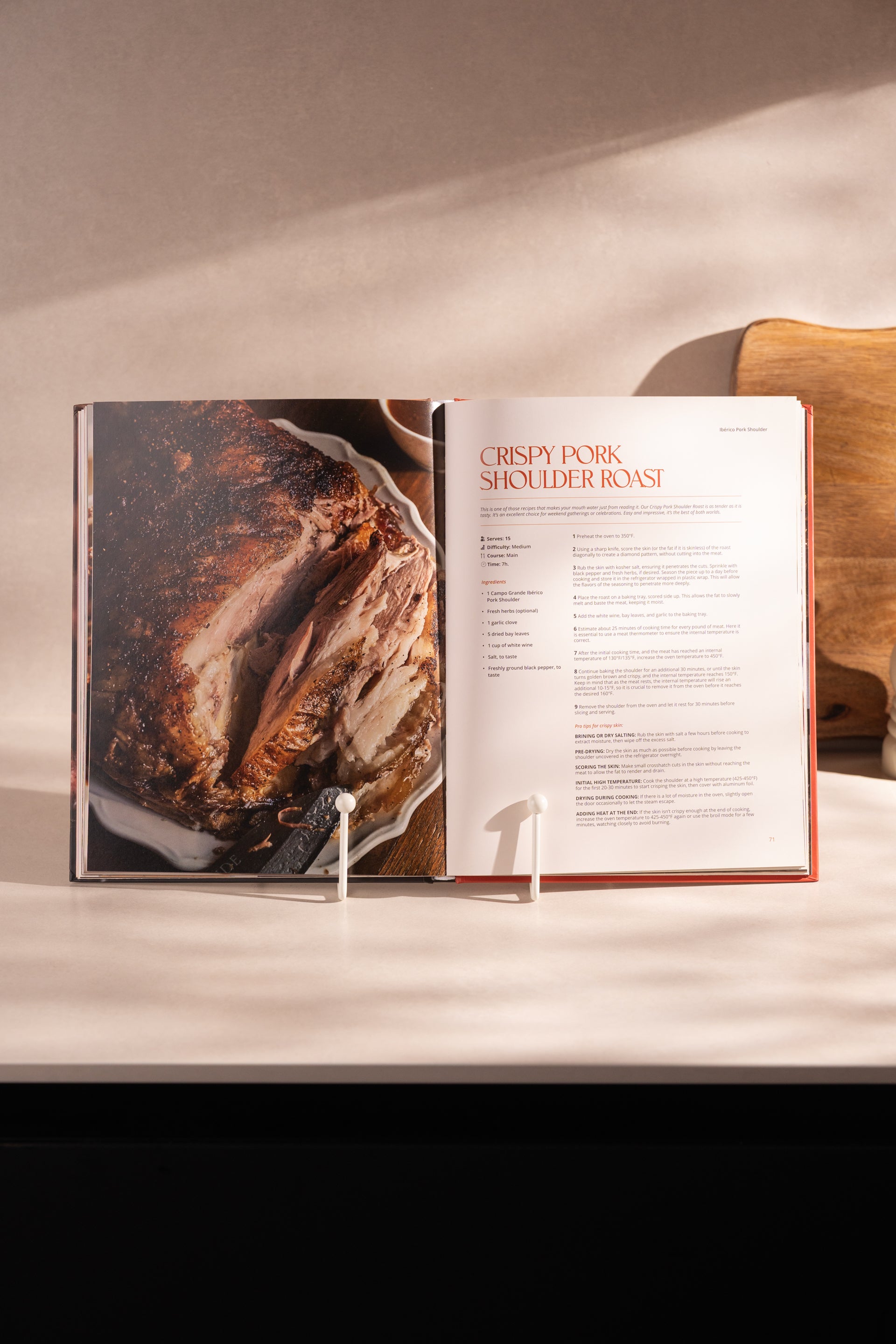Hardcover cookbook open to "Crispy Pork Shoulder Roast" recipe page with roasted meat photo on left side.