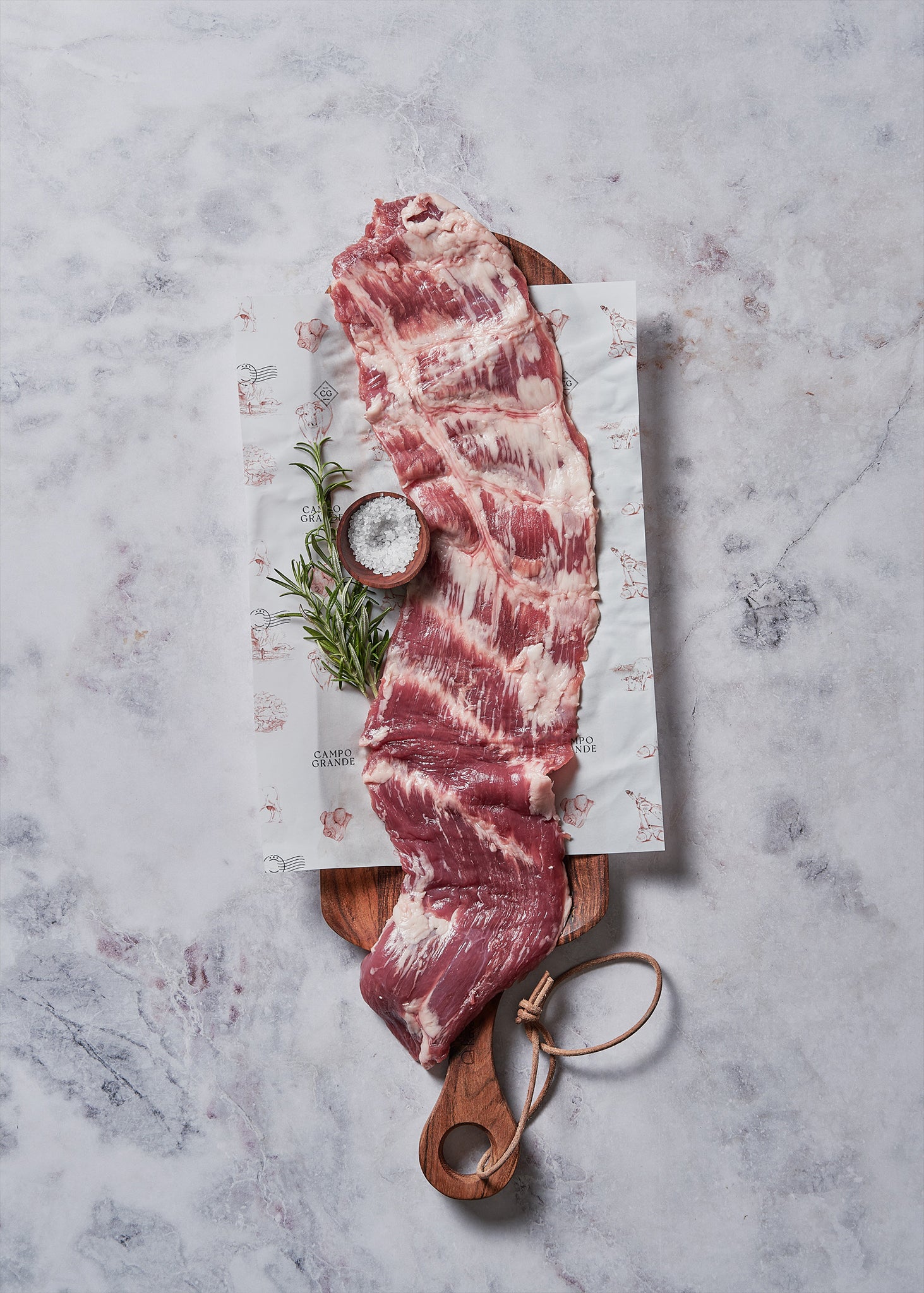 Raw ibérico flank steak with marbling displayed on wooden board with rosemary, salt, and butcher paper.