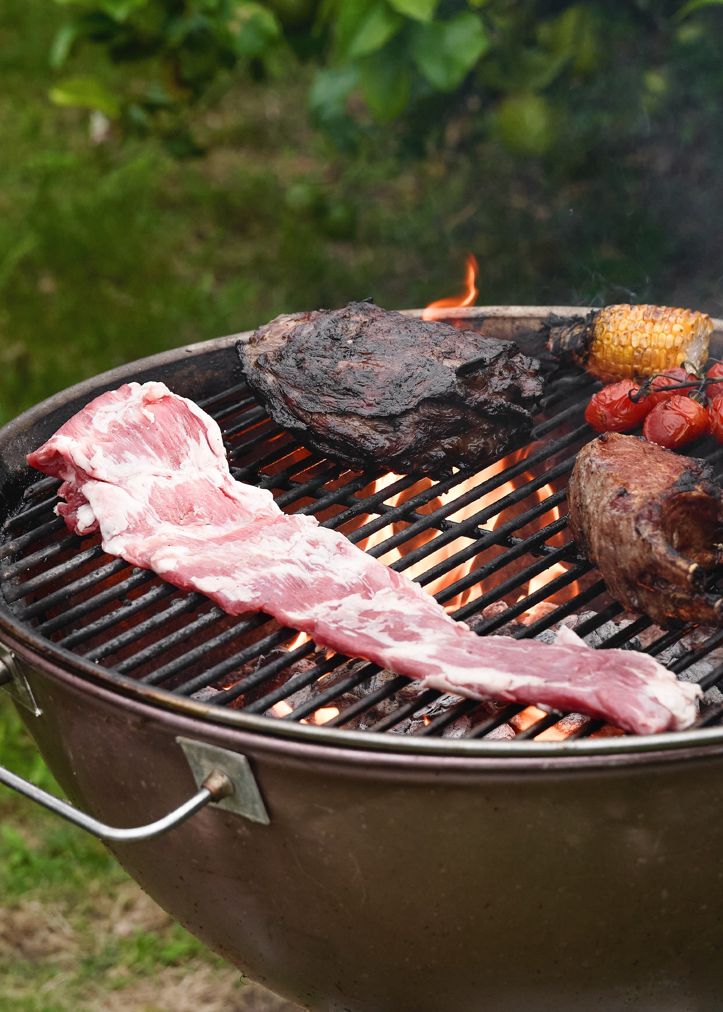 Raw flank steak with marbling on grill grate alongside charred meat, corn, and tomatoes over open flame.