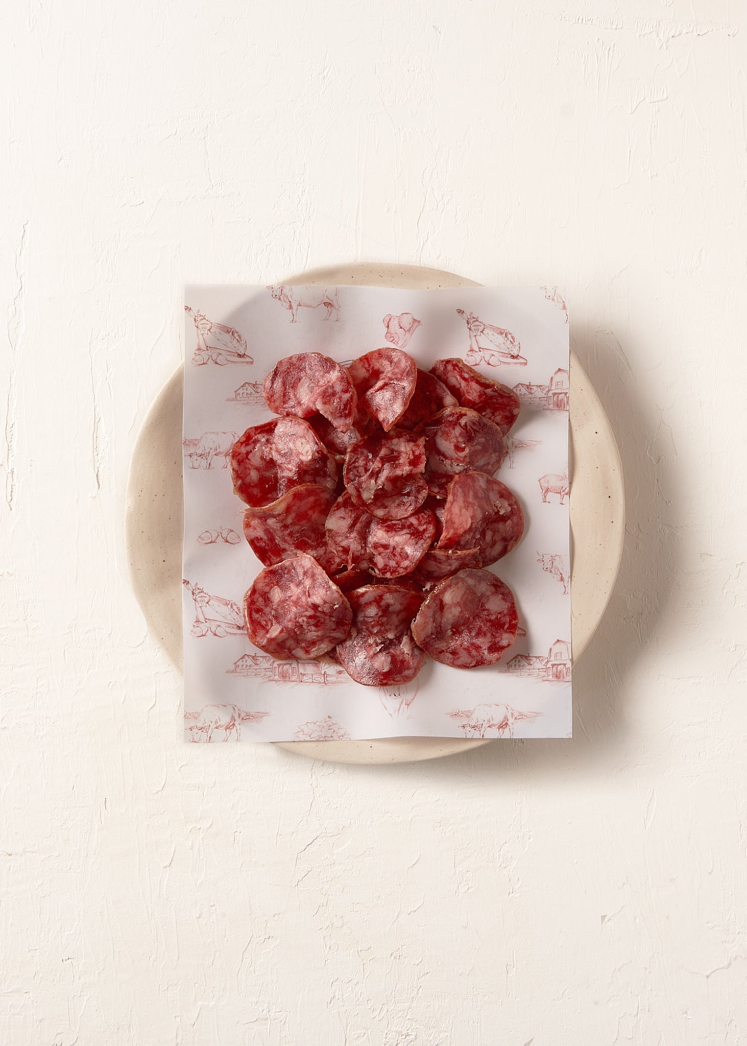Sliced red cured sausage rounds arranged on white paper with pig illustrations, placed on a cream ceramic plate.