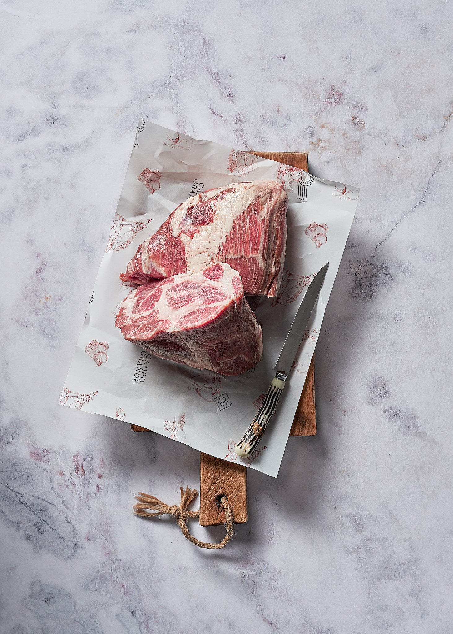 Raw red meat steaks on white butcher paper with wooden cutting board, knife, and fork on marble surface.