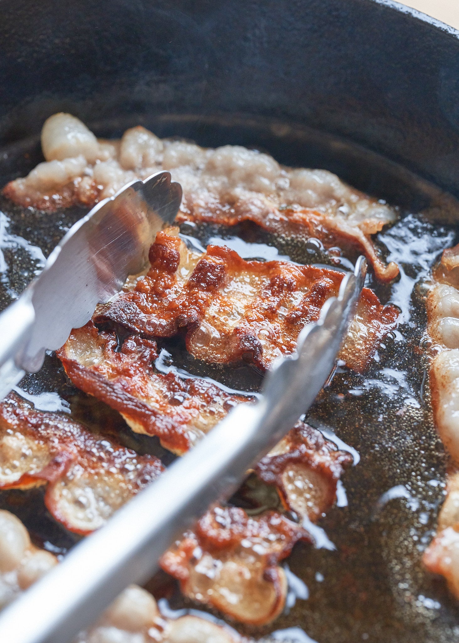 Cooked bacon strips in a dark skillet with rendered fat and tongs.
