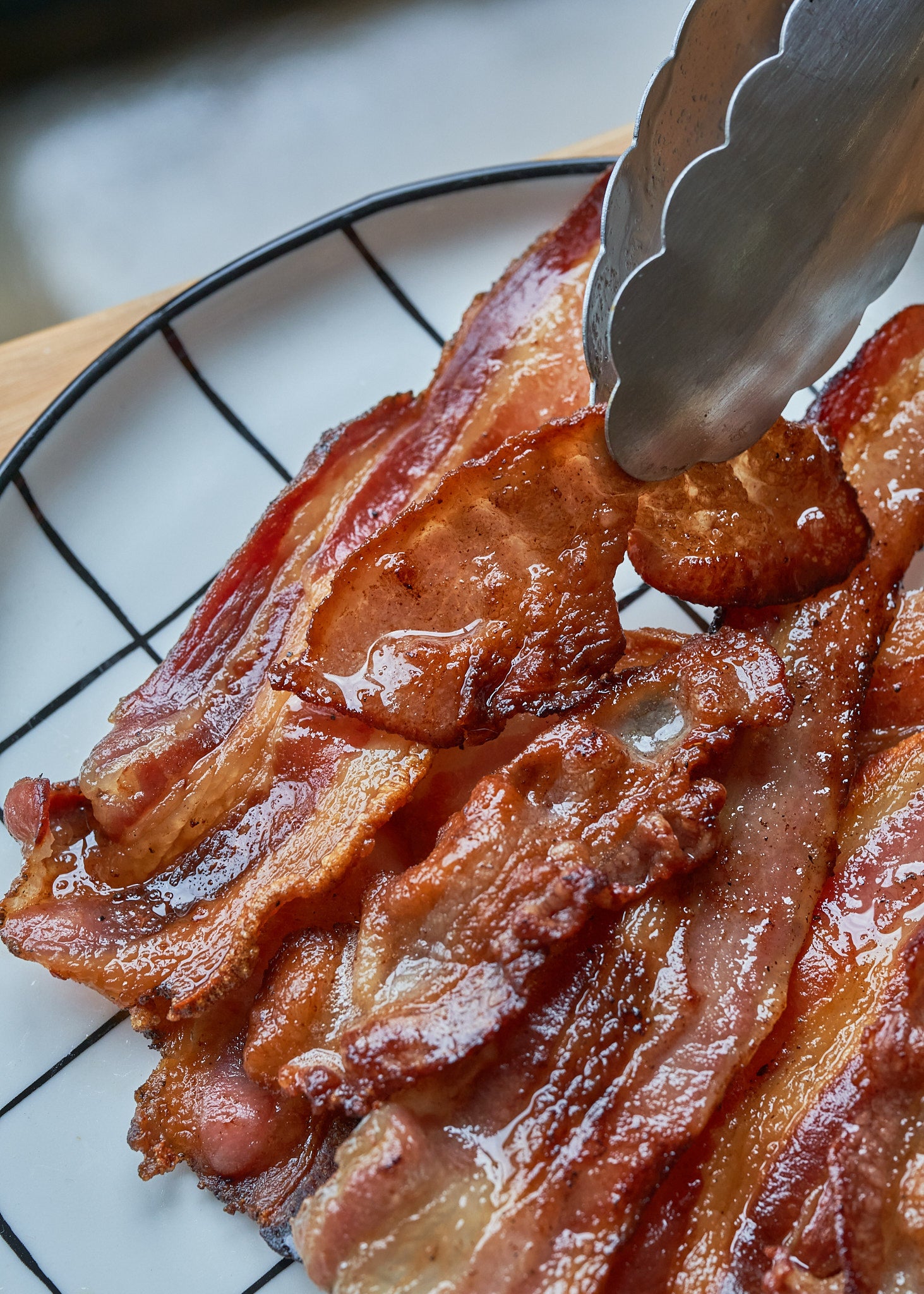 Cooked bacon strips with crispy, caramelized edges on a white plate with metal tongs.