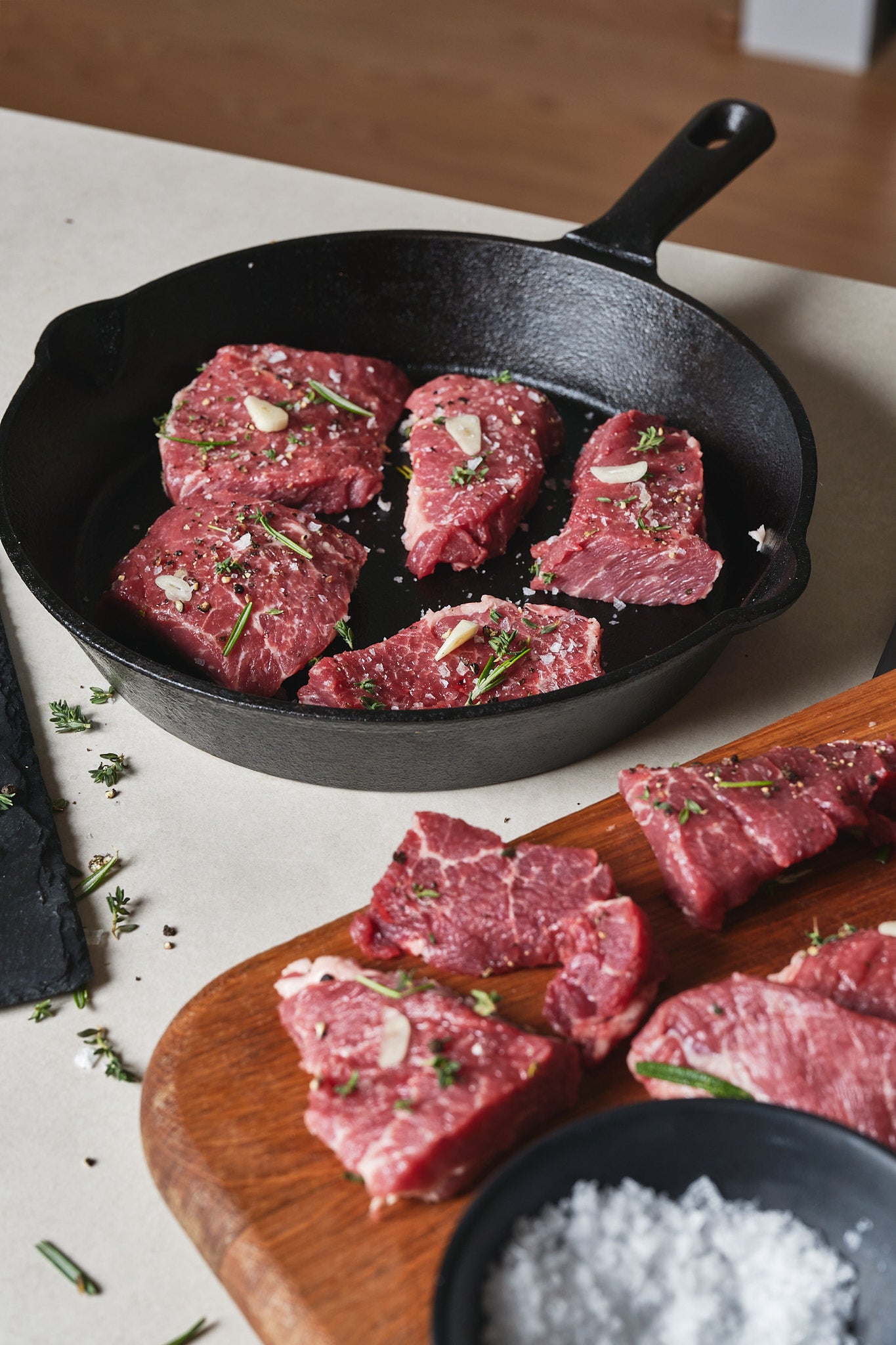 Raw Ibérico presa steaks in cast iron skillet and on wooden board, garnished with fresh rosemary and garlic slices.