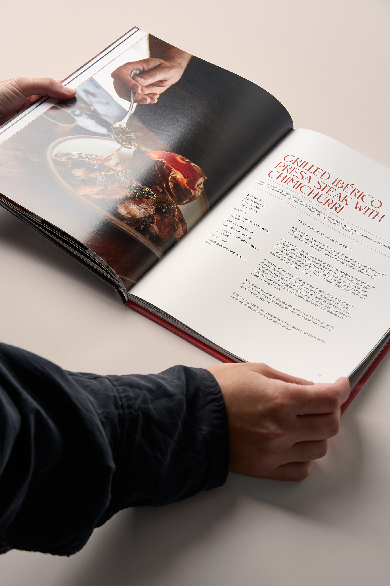 Open hardcover cookbook showing grilled Ibérico pork recipe with food photography and text instructions, held by hands.