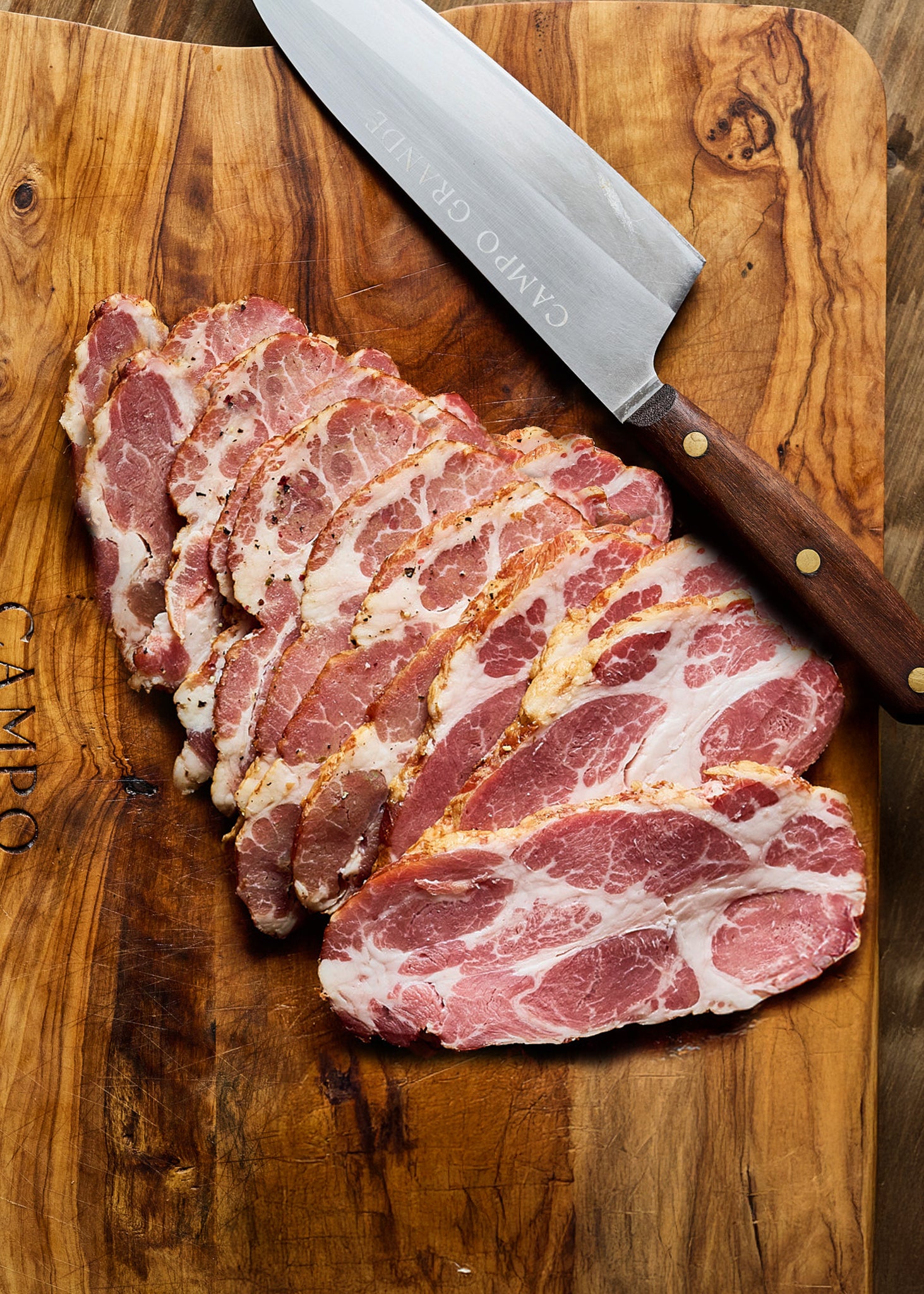 Thinly sliced cured pork with white fat marbling arranged on wooden cutting board with chef's knife.
