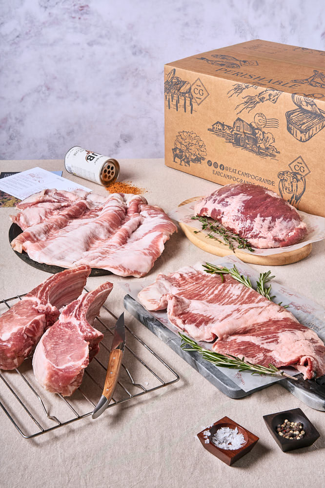 Assorted cuts of Ibérico pork on a table, including a rack of ribs, steak, and other meats, with a box labeled 'Campo Grande' in the background.