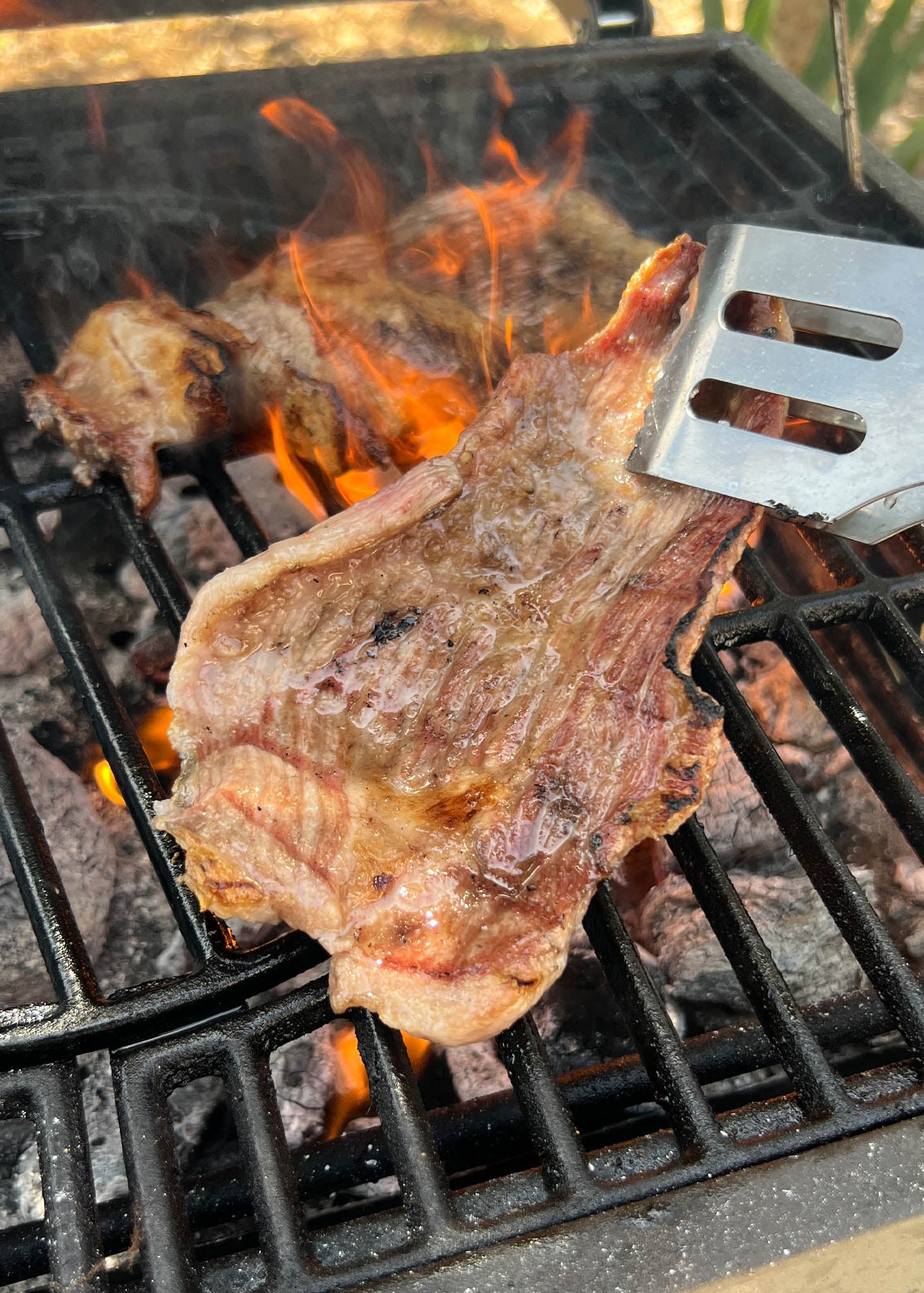 Ibérico jowl steak sizzling on a grill grate with open flames and a metal spatula nearby.