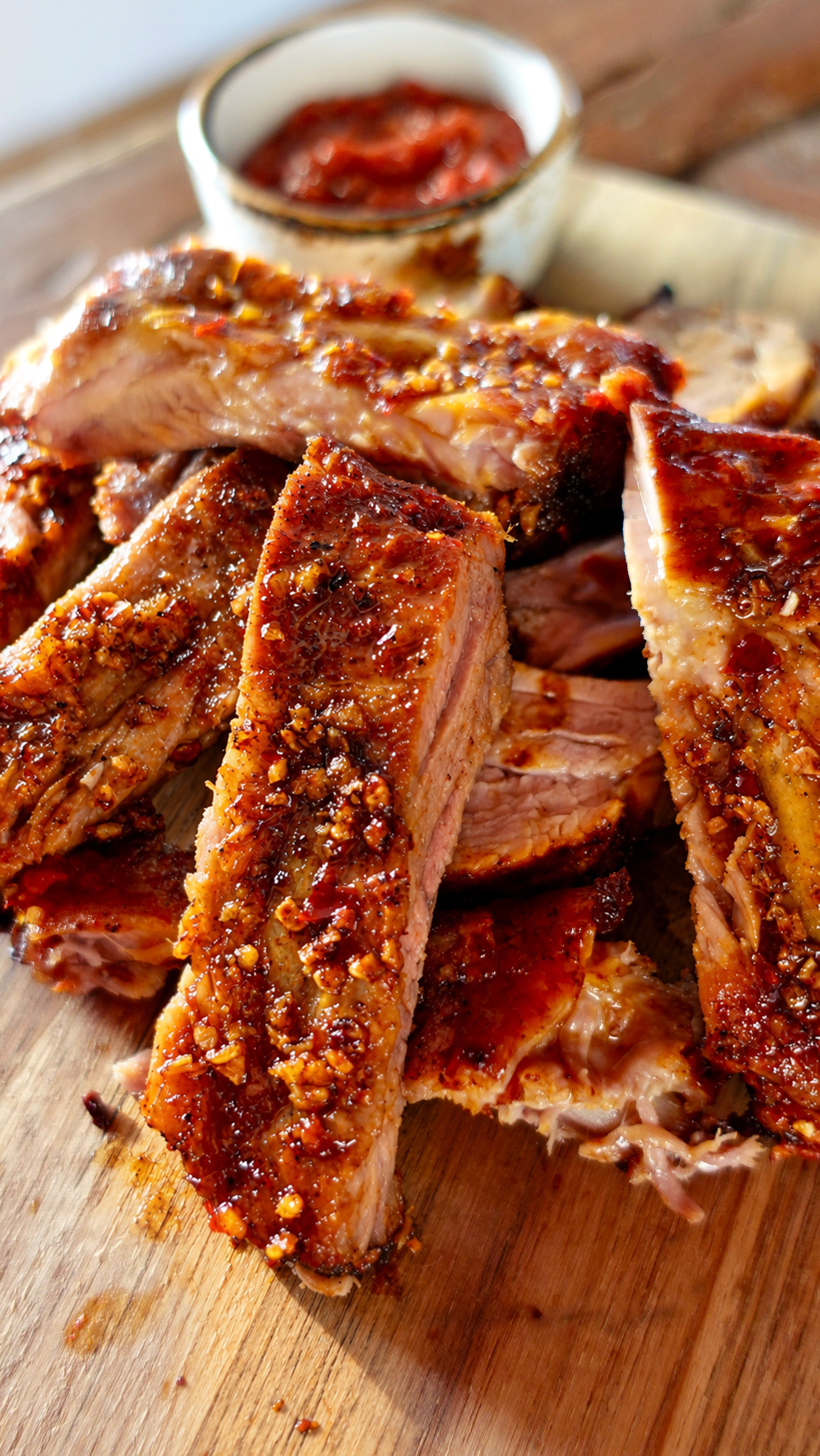Cooked ribs with caramelized glaze on wooden board next to small bowl of red sauce.