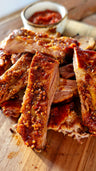 Cooked ribs with caramelized glaze on wooden board next to small bowl of red sauce.