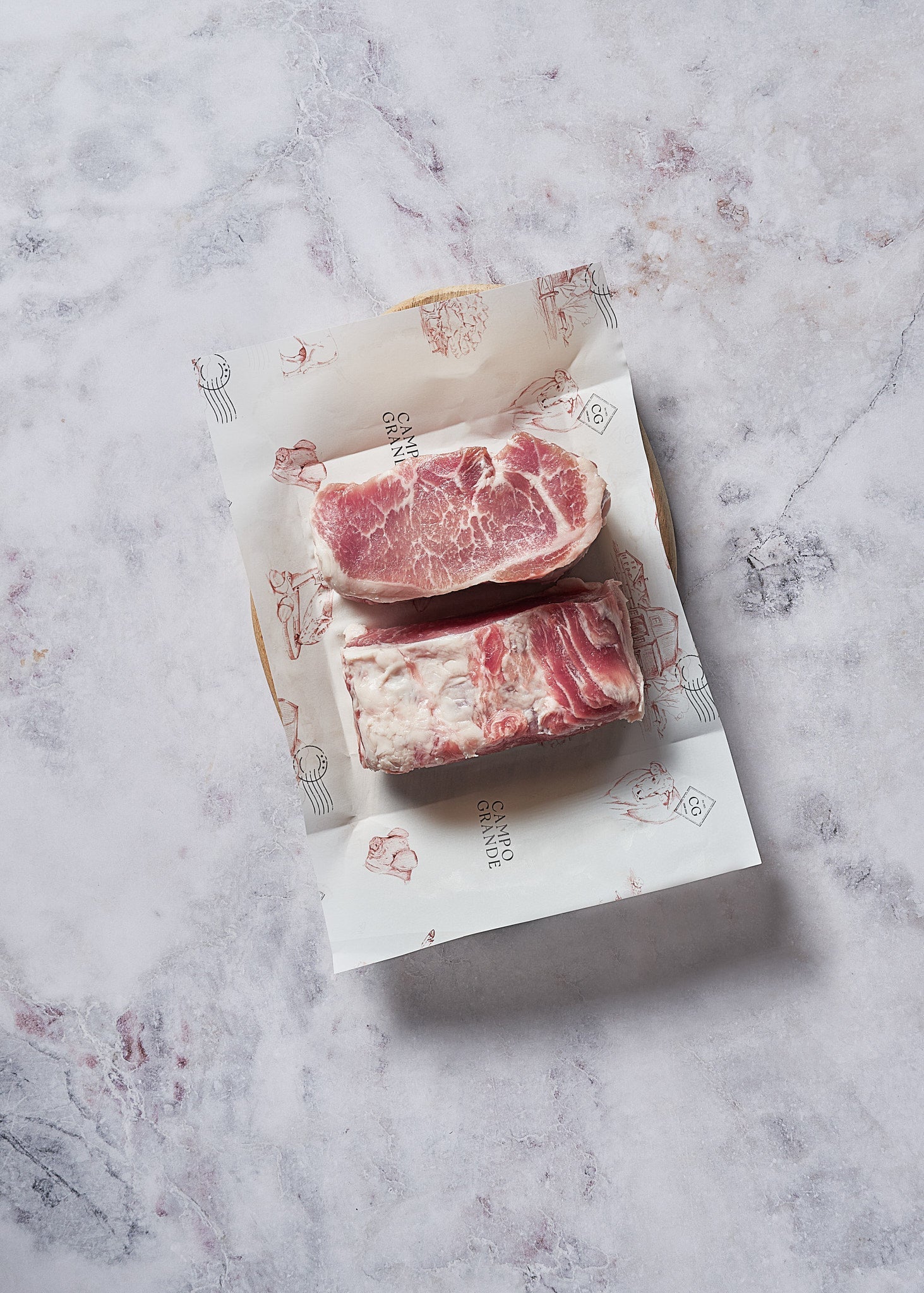 Two cuts of marbled red meat on white branded packaging with utensil illustrations, displayed on marble surface.