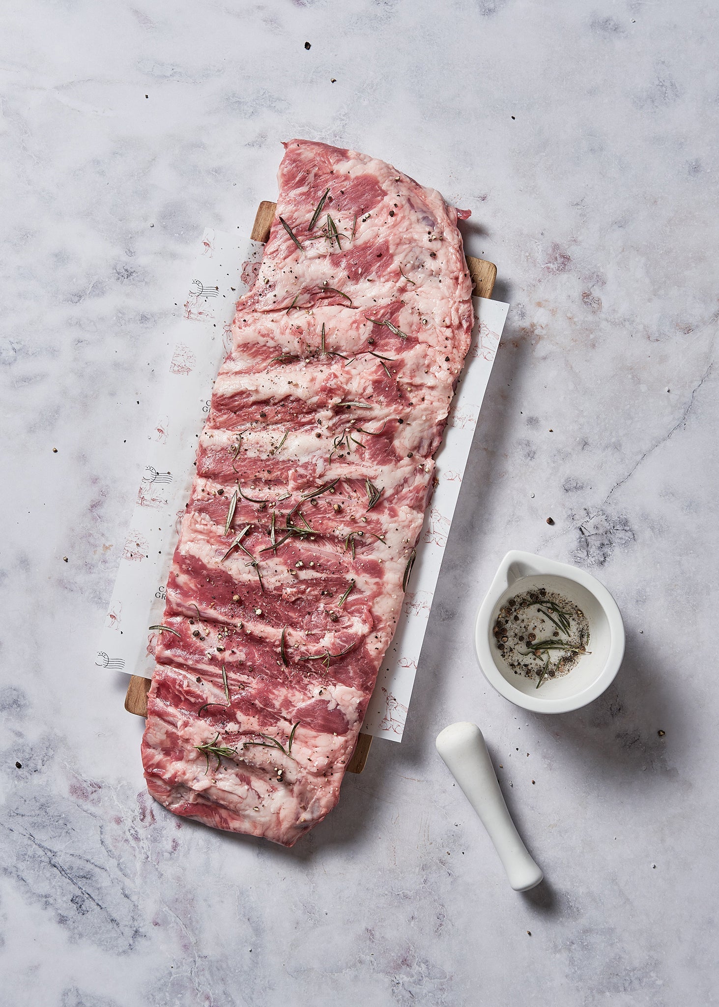 Raw meat cut with rosemary sprigs on wooden board with seasoning bowl and mortar pestle.
